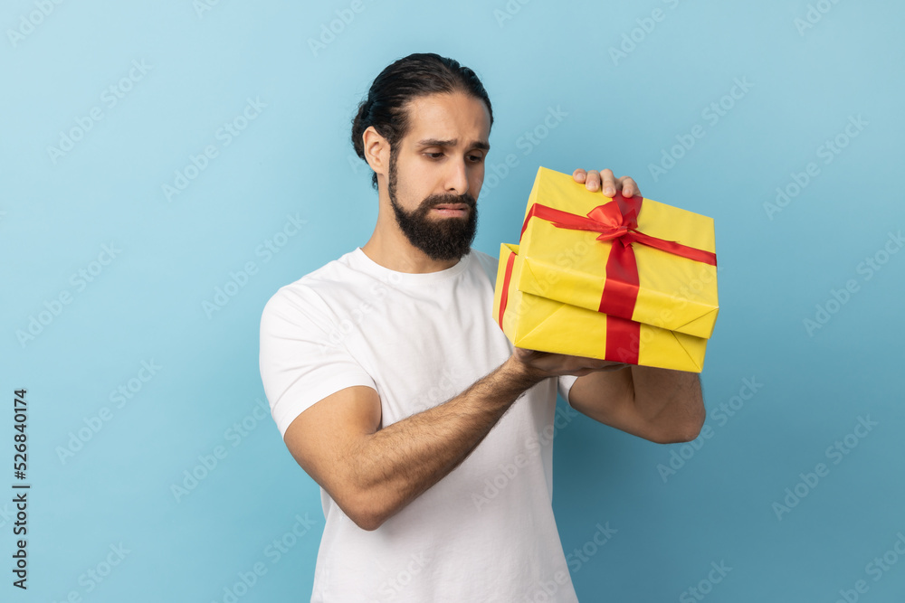 Portrait of unhappy man with beard wearing white T-shirt looking inside present box and expressing sadness, hoped to get another gift. Indoor studio shot isolated on blue background. Portrait of unhappy man with beard wearing white T-shirt looking inside present box and expressing sadness, hoped to get another gift. Indoor studio shot isolated on blue background.