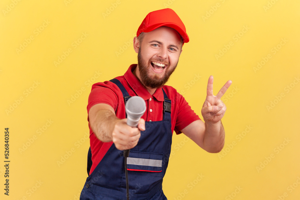 Portrait of positive bearded worker man wearing blue uniform and red cap offering microphone for answering questions and showing v sign. Indoor studio shot isolated on yellow background.