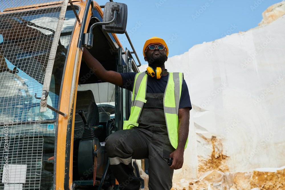 Young African American man in workwear and hardhat standing in excavator or other construction machine during work Young African American man in workwear and hardhat standing in excavator or other construction machine during work