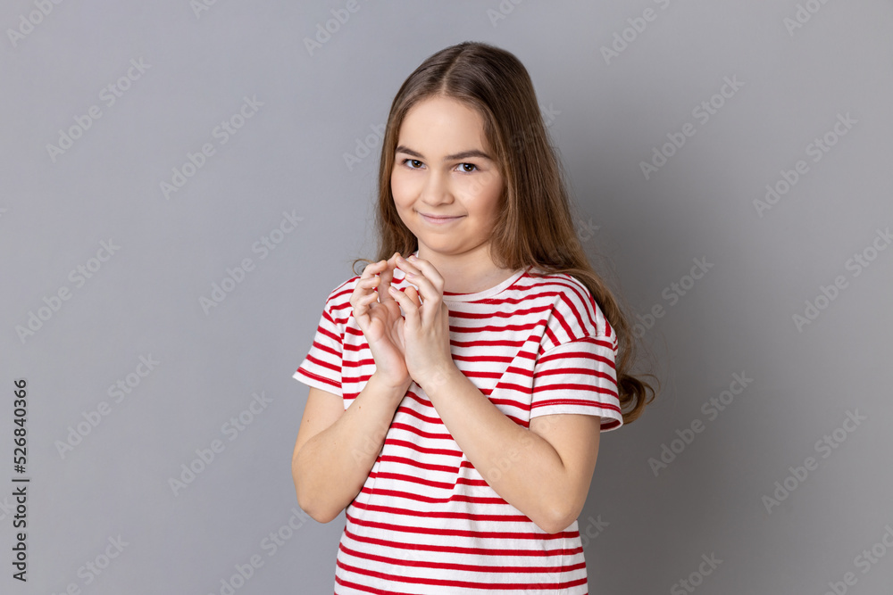 Portrait of cunning little girl wearing striped T-shirt standing looks at camera, planning something cunful, pranking somebody. Indoor studio shot isolated on gray background. Portrait of cunning little girl wearing striped T-shirt standing looks at camera, planning something cunful, pranking somebody. Indoor studio shot isolated on gray background.