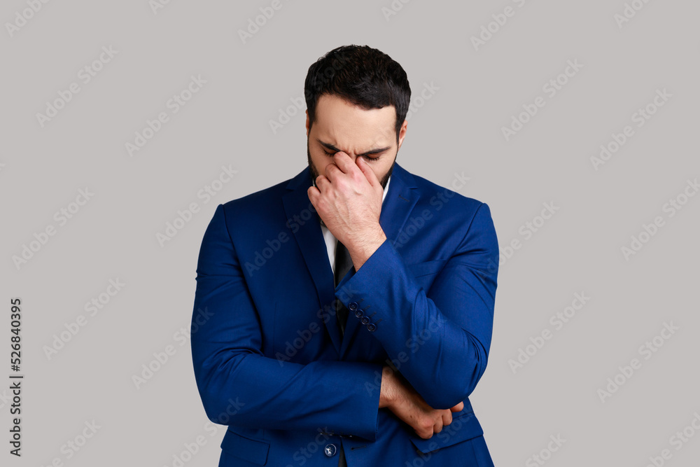 Sad upset unhappy bearded businessman hiding face in hand crying, feeling stressed worried, expressing sorrow, wearing official style suit. Indoor studio shot isolated on gray background.