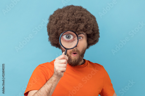 Obraz na płótnie Portrait of funny man with Afro hairstyle wearing orange T-shirt standing, holding magnifying glass and looking at camera with big zoom eye