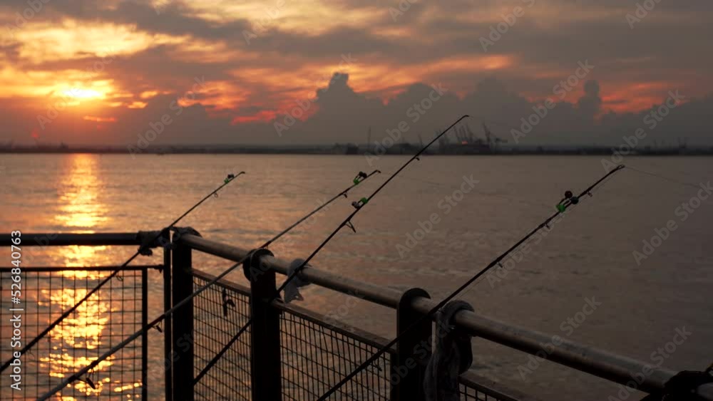 Silhouettes of a few fishing rods against the New York bay water and amazing bright sunset. The sky is yellow and orange, the waves reflect bright colors. NYC, USA. Summertime Silhouettes of a few fishing rods against the New York bay water and amazing bright sunset. The sky is yellow and orange, the waves reflect bright colors. NYC, USA. Summertime