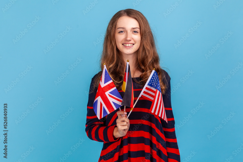 Portrait of young adult woman wearing striped casual style sweater, showing to camera British, American and German flags, expressing happiness. Indoor studio shot isolated on blue background. Portrait of young adult woman wearing striped casual style sweater, showing to camera British, American and German flags, expressing happiness. Indoor studio shot isolated on blue background.