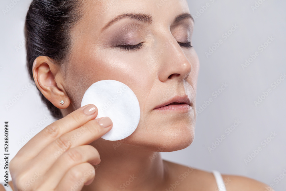 Closeup portrait of lovely beautiful middle aged woman using cotton pad with micellar water for removing make up from face, keeps eyes closed. Indoor studio shot isolated on gray background. Closeup portrait of lovely beautiful middle aged woman using cotton pad with micellar water for removing make up from face, keeps eyes closed. Indoor studio shot isolated on gray background.