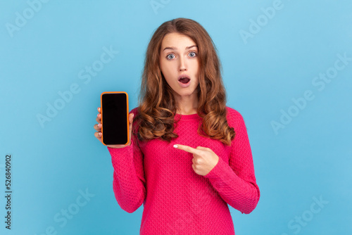 Shocked amazed woman with widely open mouth wearing pink pullover, holding smart phone in hands with blank display for advertisemant. Indoor studio shot isolated on blue background. photo