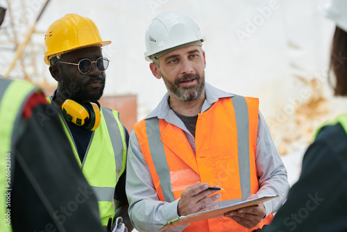 Confident foreman with document and pen explaining something to subordinates or giving instructions while looking at one of them