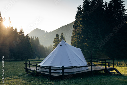 glamping tent in green meadow surrounded by fir tree forest at sunrise