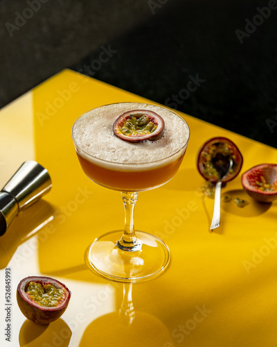 Glass of Porn Star Martini cocktail with passionfruit on yellow table with dark background, raw fruits around. Summer exotic fruit alcohol refreshing drink with foam. Copy space.