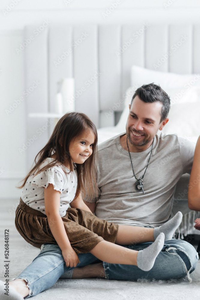 Obraz premium Little cheerful daughter sits on her father and smiles. Happy dad and his little daughter have fun at home. Good relationship between father and daughter. Girl dressed in a skirt and t-shirt
