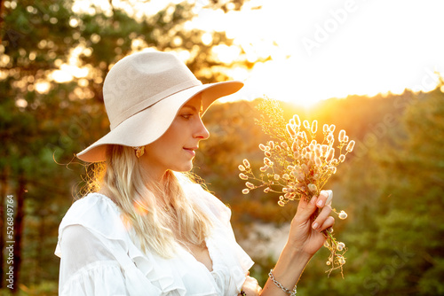 Εκτύπωση καμβά Side view of young beautiful woman standing at sunset in forest, holding, looking at bouquet of wildflowers in summer