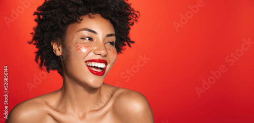Laughing smiling african american girl with bare shoulders, glowing body, curly hair, looking aside at empty space on red background