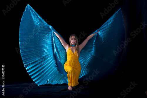 mujer joven vestida de amarillo posando con alas de Isis de color azul , con fondo negro y luces de fuego
