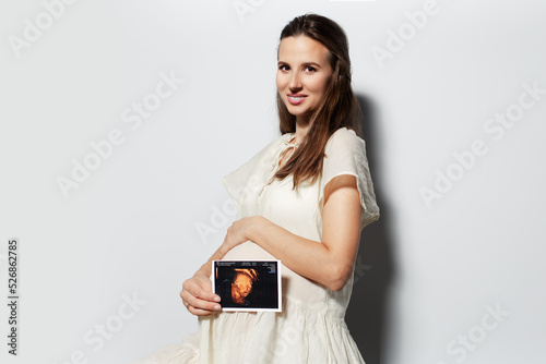Studio portrait of young happy pregnant woman touching the belly, showing ultrasound scan on white background.