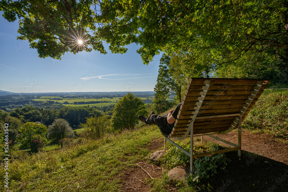 Sonnenbank mit Aussicht nahe der Burg Hohenzollern auf der schwäbischen Alb, ein nicht erkennbarer Wanderer entspannt sich auf der Bank