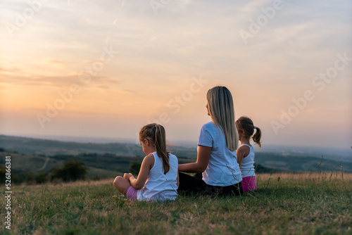 Mother and daughters doing yoga exercises on grass in the park at the sunset. Family having fun outdoors. Concept of friendly family and of summer vacation.
