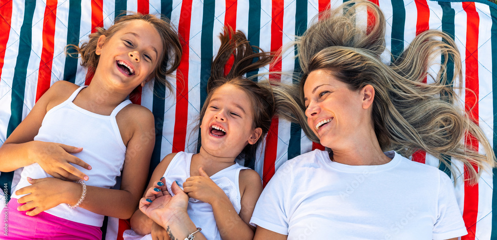 Fototapeta premium Happy mother and daughters smiling and laughing lying on blanket at the lawn in summer park. Family spend their free time together on vacation.