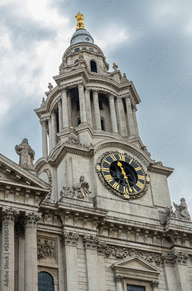 London, England, UK - July 6, 2022: St. Paul's Cathedral. Closeup of SW tower spire with golden top and clock. Stone statues on corners and above under gray cloudscape.
