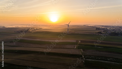 Colorful sunrise over panoramic farmfields with foggy horizon and huge wind turbine. Bright sun glare. Environmental awareness. Horizontal shot. High quality photo