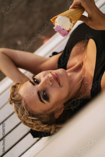 Beautiful mature woman eating ice cream in a waffle cone while lying on a park bench.