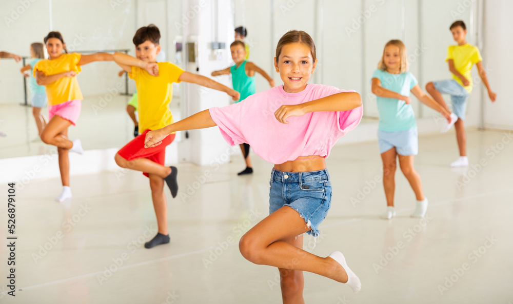 Portrait of cheerful preteen girl learning energetic dance moves with group of children in choreography class .