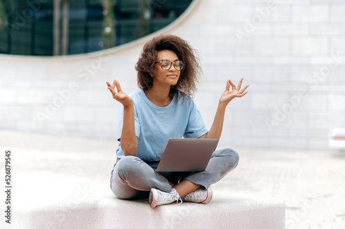 Calm relaxed curly african american young woman with glasses, office worker, sits in a lotus position outdoors near the business center, meditates, relaxes, closed her eyes. Break during online work