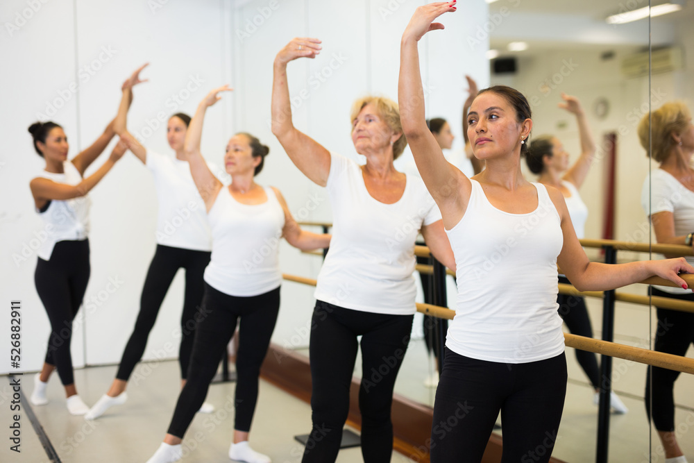 Women of different ages standing in row and training ballet moves. Trainer correcting them.