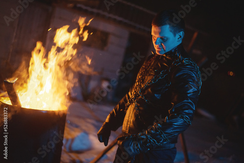 Motorbiker in the black leather jacket with the spikes in the blue light on the burning fire background.