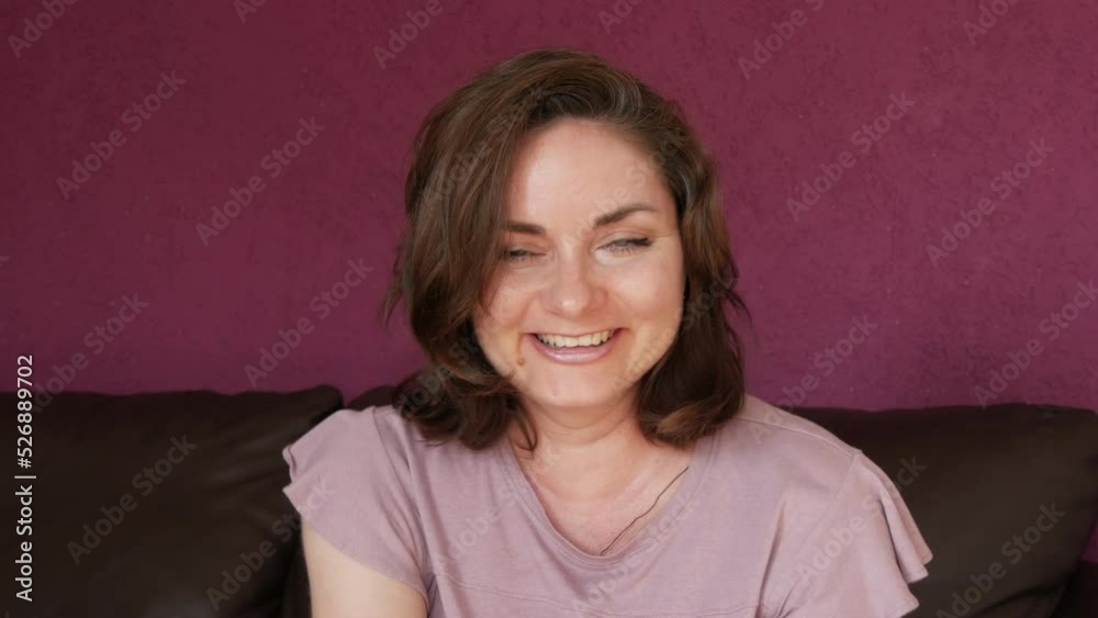 Close-up portrait of a young beautiful woman with green eyes and dark hair, who looks directly into the camera fooling around, grimacing having fun and dancing on a leather sofa in the room
