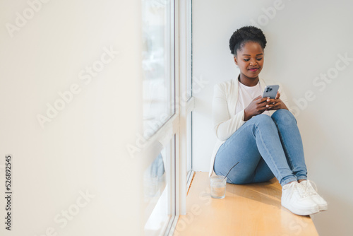 Smiling African girl sitting on a cafe windowsill checking the data on the mobile phone. Afro Hairstyle, enjoying moment, happy people, city life © Strelciuc