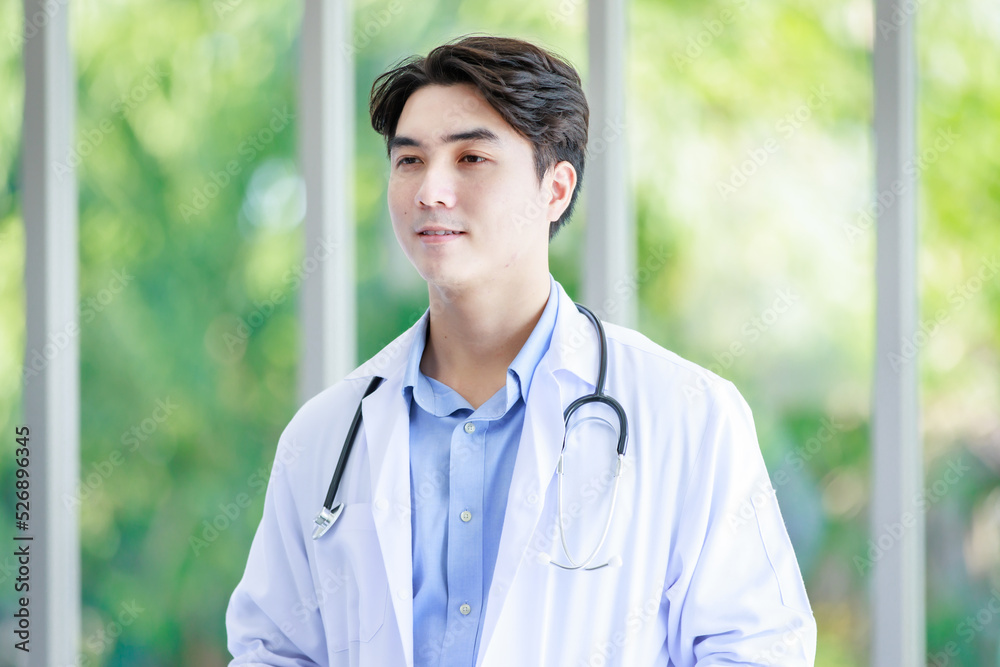 Closeup shot of Asian young cheerful happy successful professional male intern practitioner doctor in white lab coat with stethoscope standing smiling alone in hospital hallway on blurred background Closeup shot of Asian young cheerful happy successful professional male intern practitioner doctor in white lab coat with stethoscope standing smiling alone in hospital hallway on blurred background