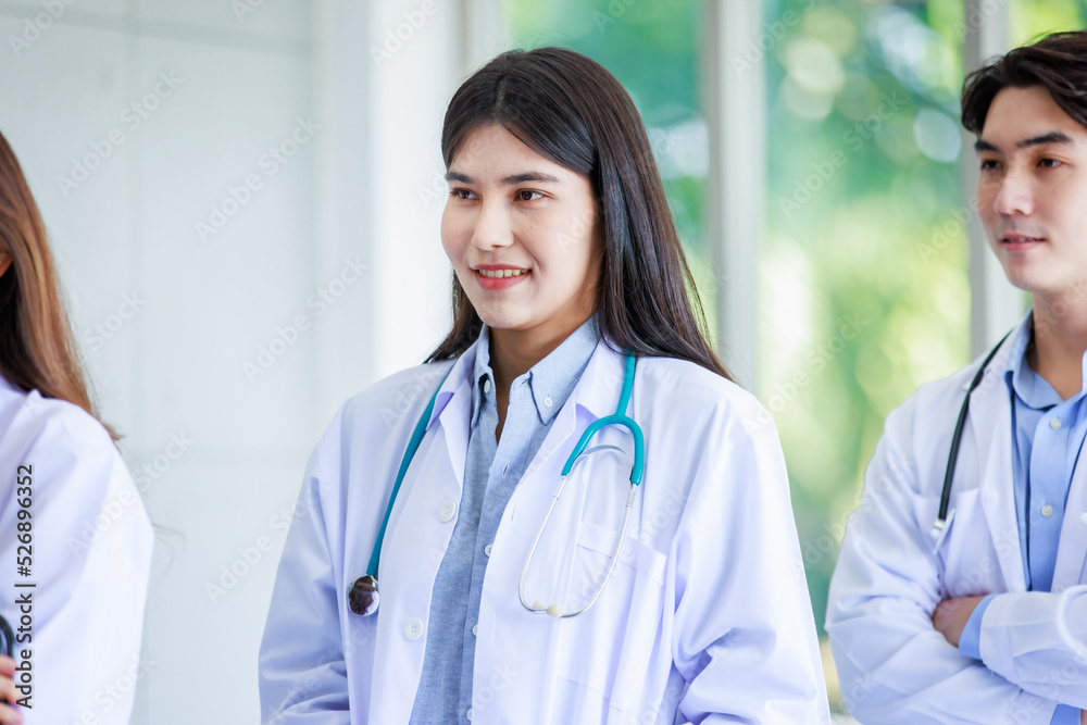 Closeup shot of Asian young cheerful happy professional intern practitioner doctor in white lab coat with stethoscope standing smiling in front of male and female colleagues on blurred background Closeup shot of Asian young cheerful happy professional intern practitioner doctor in white lab coat with stethoscope standing smiling in front of male and female colleagues on blurred background