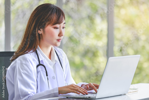 Asian successful professional young female doctor in white lab coat uniform with stethoscope sitting smiling using typing laptop notebook computer working researching online in hospital clinic office