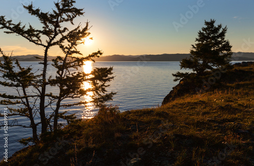 Baikal Lake early summer morning. Silhouettes of beautiful larch trees on the shore of the Small Sea against the backdrop of the rising sun over the water. Scenic landscape. Natural seaside background