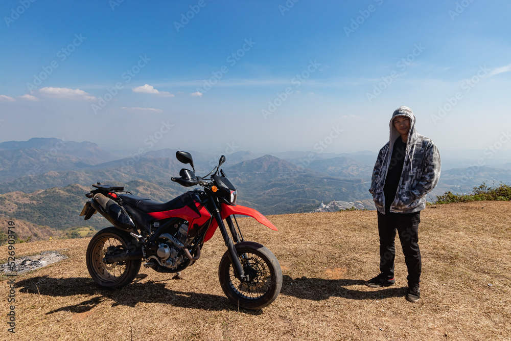 A motorcyclist is standing beside a motorcycle at the top of the mountain. In the distant background are roads and temples. Pha tud, Phetchabun Province, Thailand A motorcyclist is standing beside a motorcycle at the top of the mountain. In the distant background are roads and temples. Pha tud, Phetchabun Province, Thailand