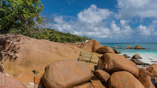A picturesque beach on a tropical island. Lush green vegetation on the hillside. Clouds in the blue sky. The turquoise ocean is calm. In the foreground is a pile of granite boulders. Seychelles