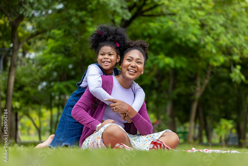African American mother is playing piggyback riding and hugging with her young daughter while having a summer picnic in the public park for wellbeing and happiness concept