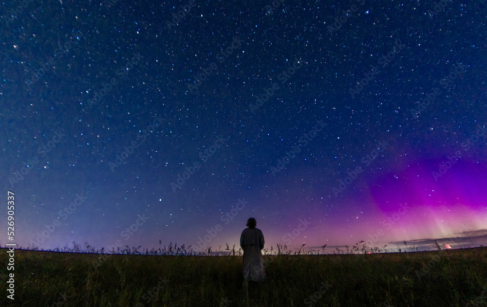 Fototapeta premium abstract night photo of a figure of a lonely person in a field under a starry sky and northern lights, starfall. Astrophotography