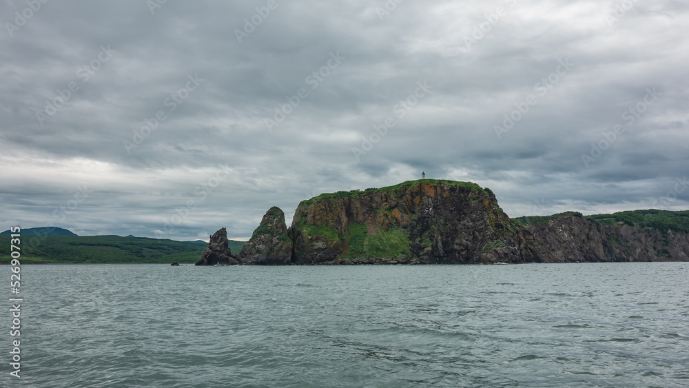 The harsh coast of Kamchatka against the background of a cloudy sky and the ocean. Sheer cliffs and green hills. Avacha Bay