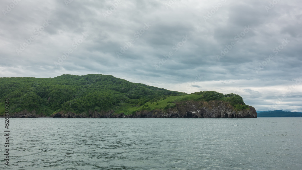 Green hills and steep cliffs of the Kamchatka coast against a background of cloudy sky and ocean. Avacha Bay Green hills and steep cliffs of the Kamchatka coast against a background of cloudy sky and ocean. Avacha Bay