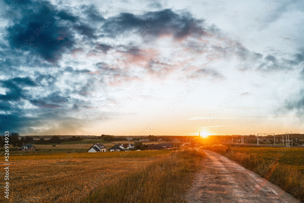 Fototapeta premium Rural country road at the beautiful summer sunset nature landscape. Empty road and tranquil evening scene with warm sunlight. The dramatic sky above the farm.