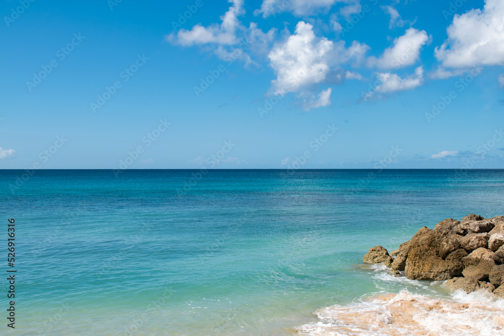 A view of a bunch of rocks on a Caribbean beach in Barbados. There is calm turquoise water and a cloudy blue sky.