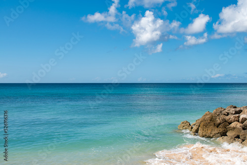 A view of a bunch of rocks on a Caribbean beach in Barbados. There is calm turquoise water and a cloudy blue sky.