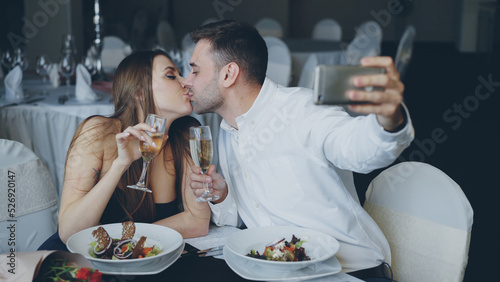 Obraz na plátně Attractive loving couple is taking selfie with champagne glasses using smartphone while having romantic dinner in restaurant