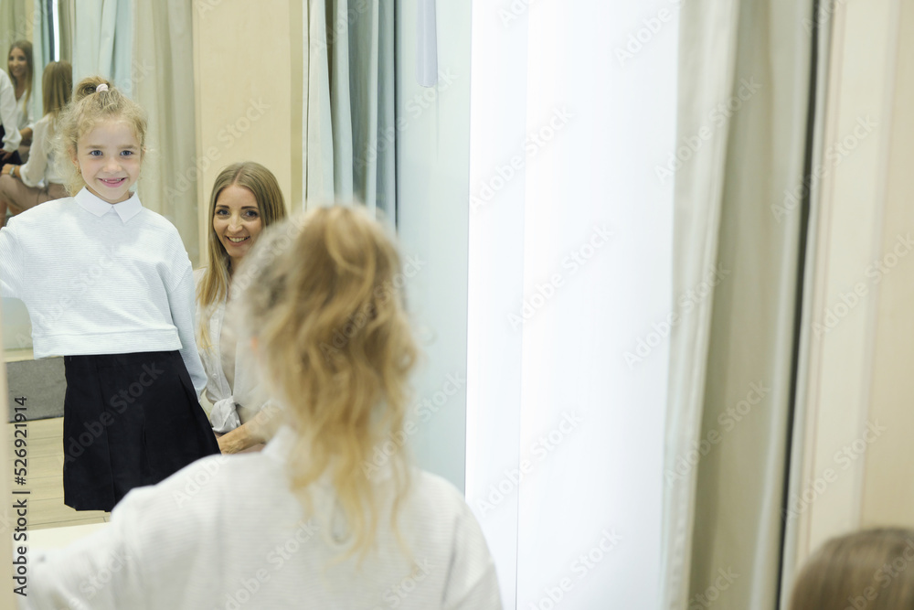 Fototapeta premium Mother and her little daughter in a fitting room in a shopping mall. The girl tries on a jacket and a skirt before buying, the child looks at himself in the mirror and smiles