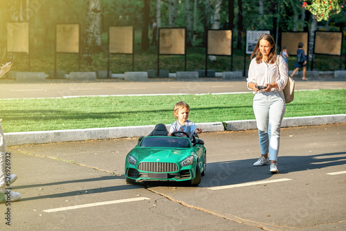 Mom controls children car with remote at walk with toddler child in public park. Toddler boy sits in green car holding steering wheel looking aside