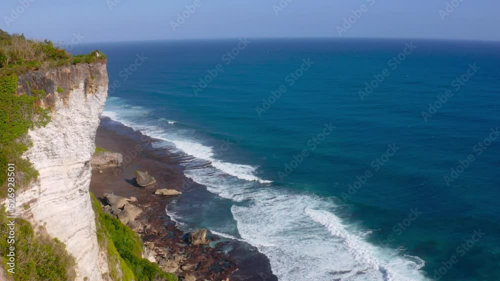 Majestic cliff standing tall beside coastline of beach and low tides moving towards them. Aerial.