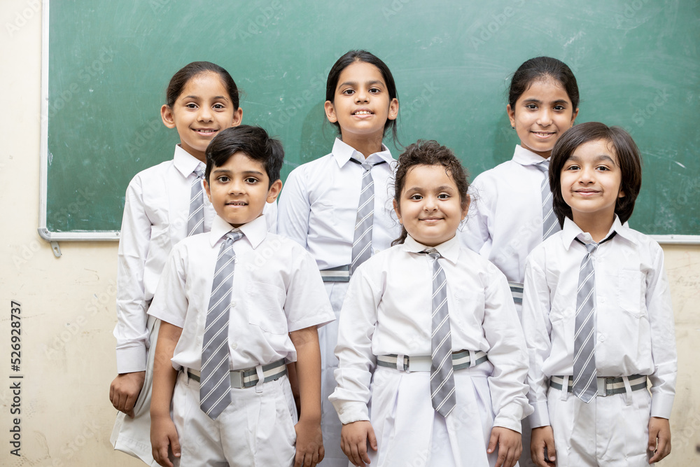 Portrait of happy indian school kids in uniform standing in classroom with chalk board in the background, Elementary school, Education concept.