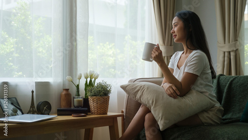 Relaxed young woman sitting on sofa and drinking hot coffee, enjoying weekend at home.
