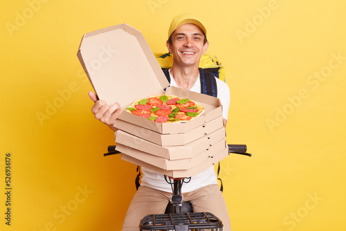 Indoor shot of postive happy courier wearing cap holding open pizza box from restaurant, riding bicycle, expressing optimistic emotions, posing isolated on yellow background photo